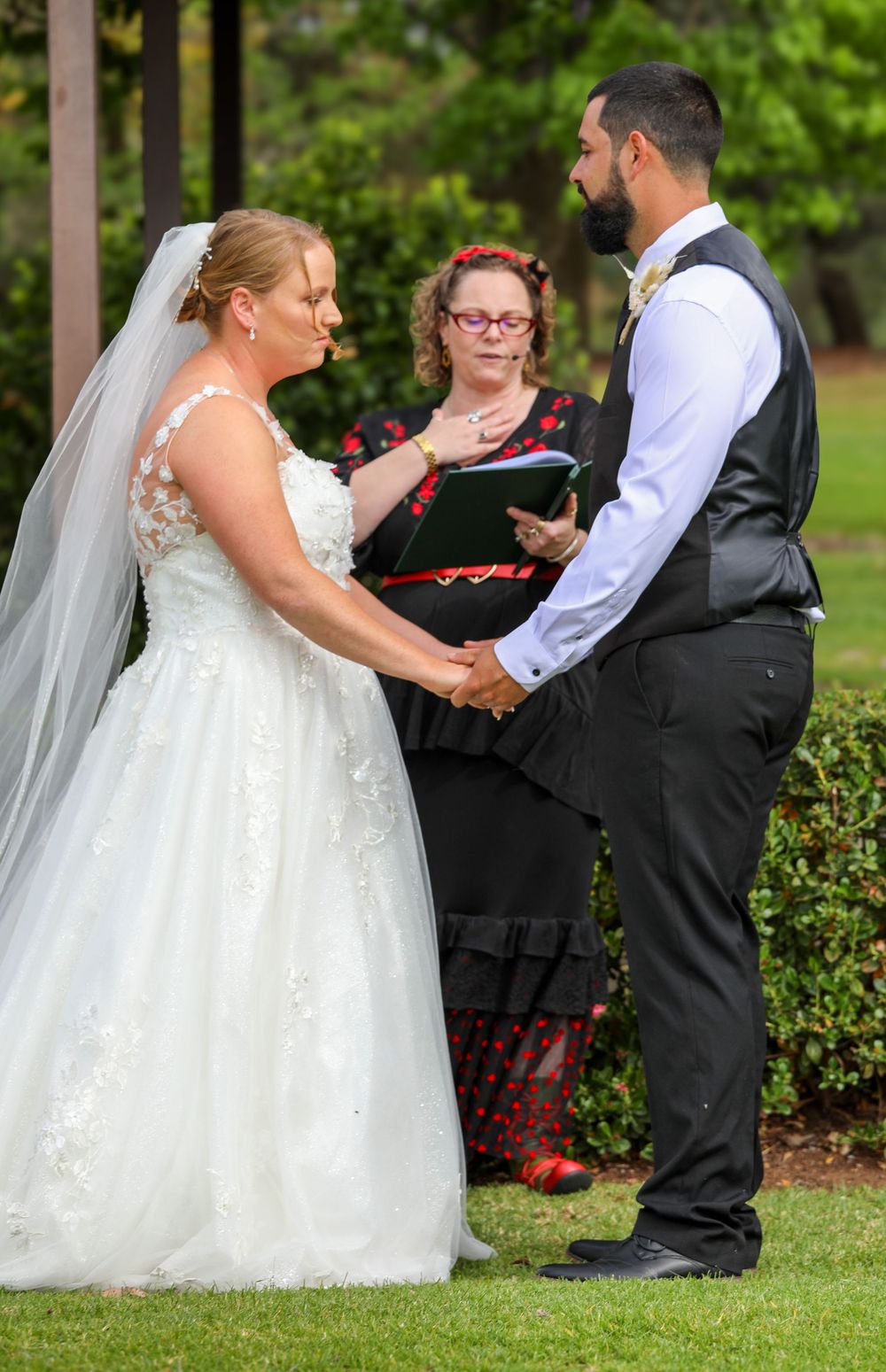 An outdoor wedding ceremony takes place on a lawn with decorative hedges and greenery.