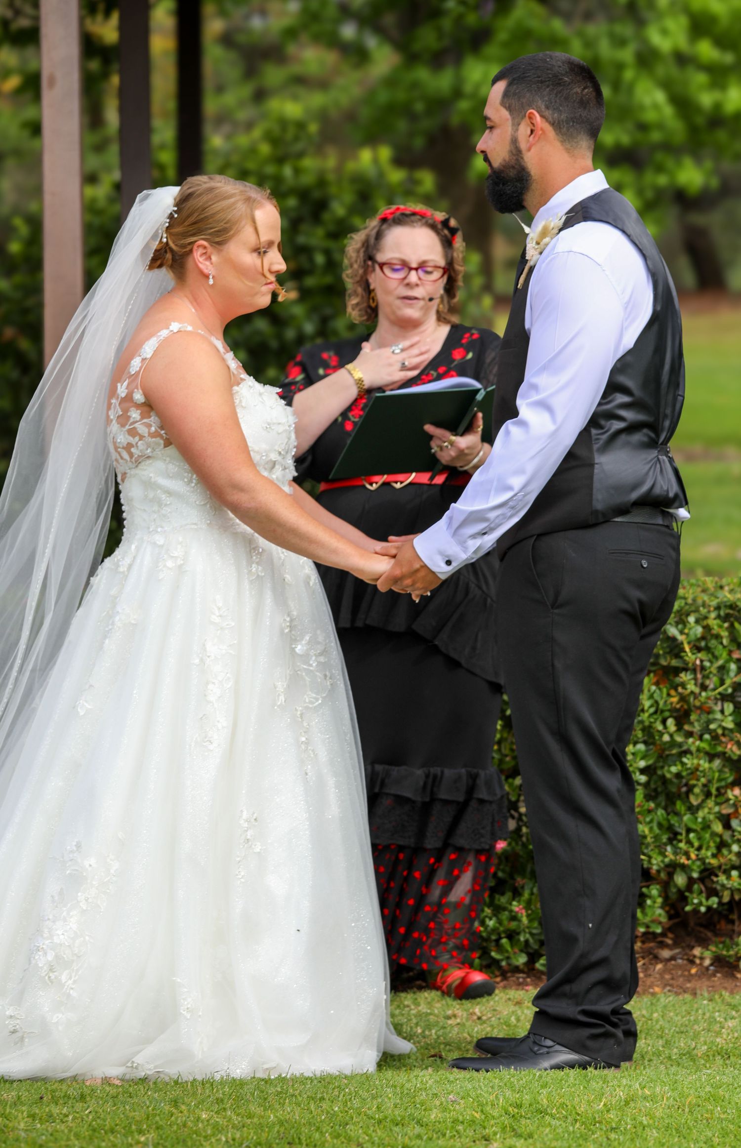 An outdoor wedding ceremony takes place on a lawn with decorative hedges and greenery.