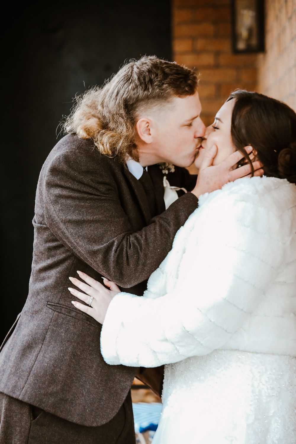 A tender kiss shared between newlyweds against a brick wall background.