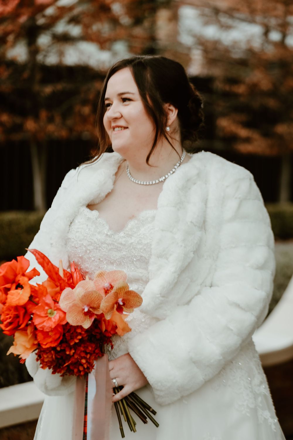 A person in a white fur wedding coat holds vibrant coral and red flowers.