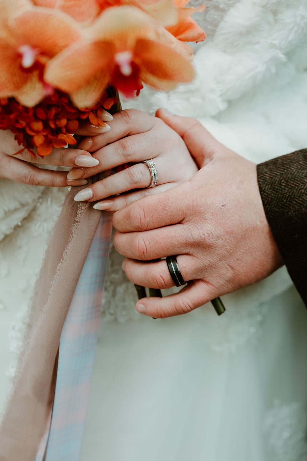Wedding rings and hands holding a coral-colored bouquet against white fabric.