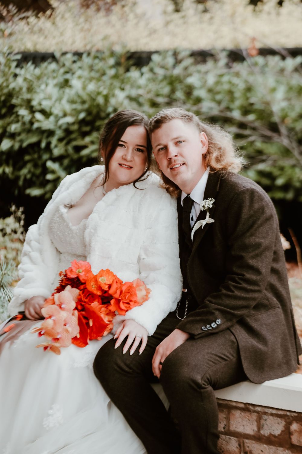 A romantic couple embraces on a bench with coral-colored flowers.