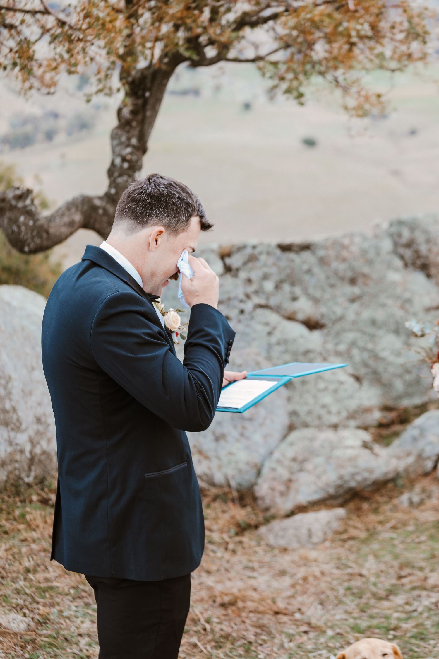 A wedding guest in black suit wipes tears during an emotional ceremony outdoors.