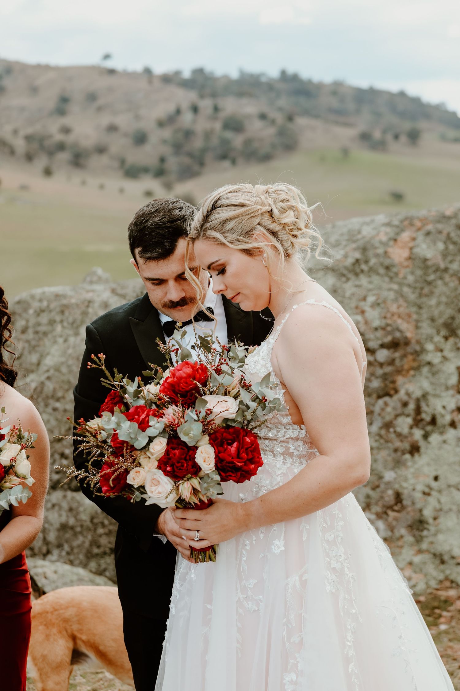 A couple admires a vibrant red and white wedding bouquet in a rustic outdoor setting.
