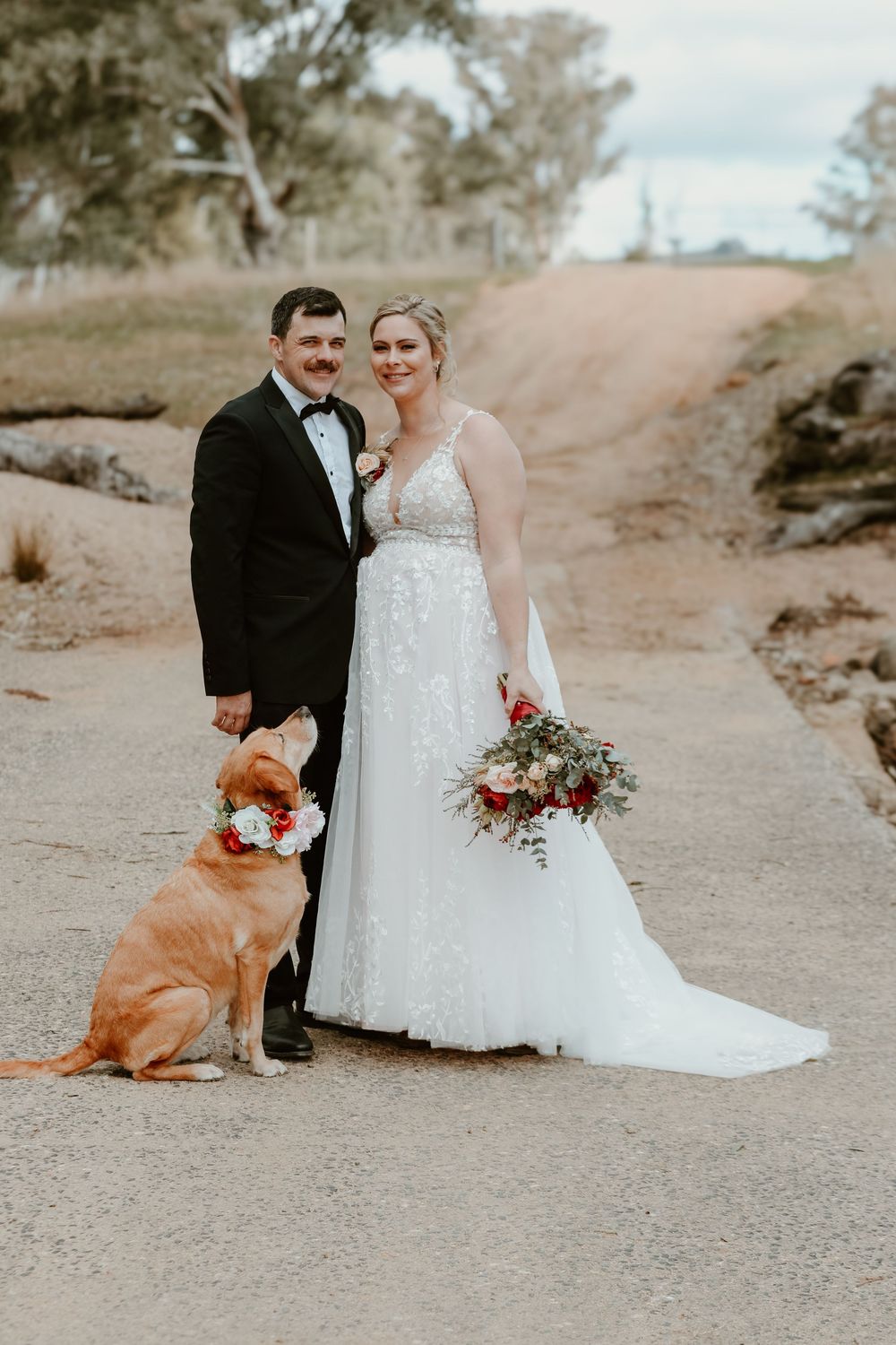 A wedding couple poses with their golden retriever dog on a desert path at sunset.