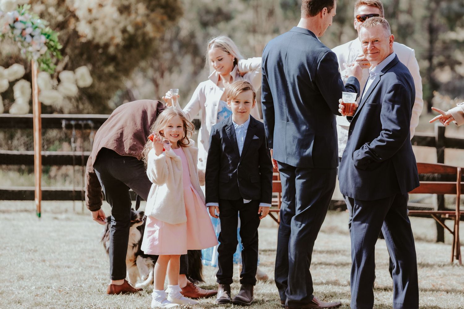 Wedding guests gathered at an outdoor rustic venue with wooden fencing.