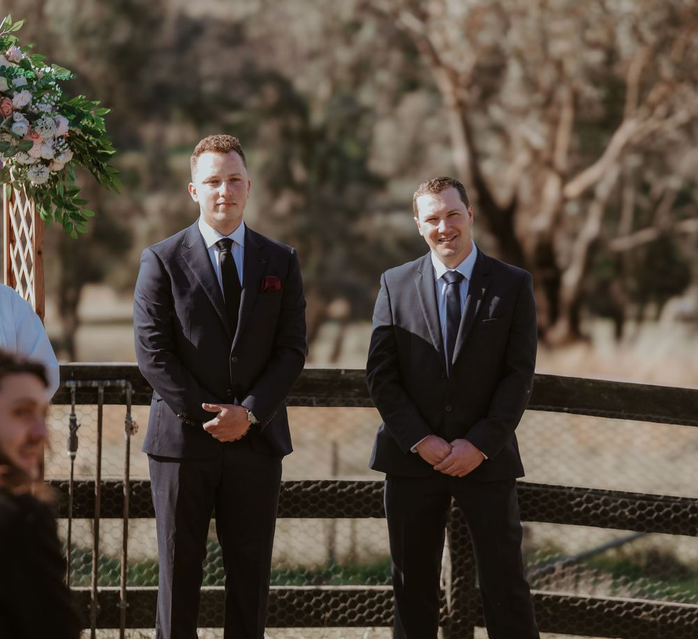 Two people in suits stand next to dark wicker furniture at an outdoor wedding ceremony location.
