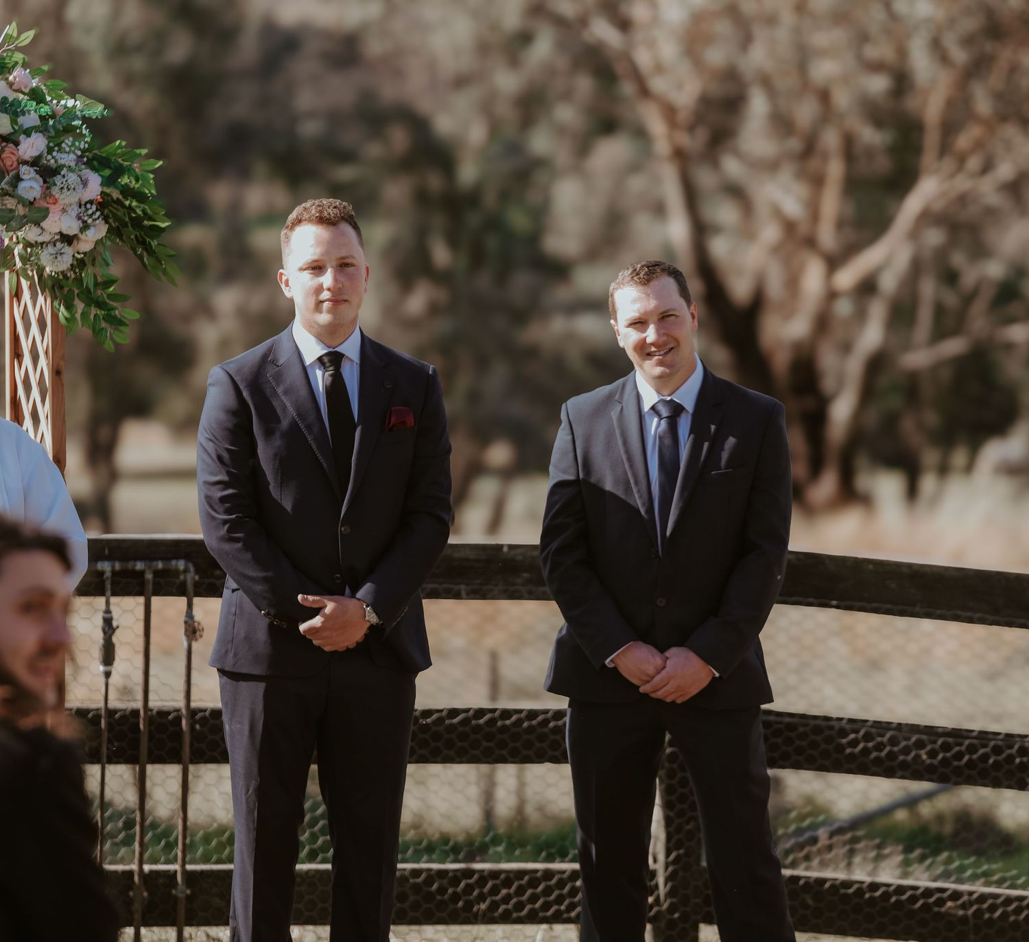Two people in suits stand next to dark wicker furniture at an outdoor wedding ceremony location.