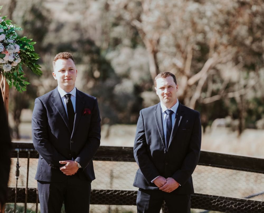 Two people in formal navy suits stand on an outdoor stone pathway during a wedding photoshoot.