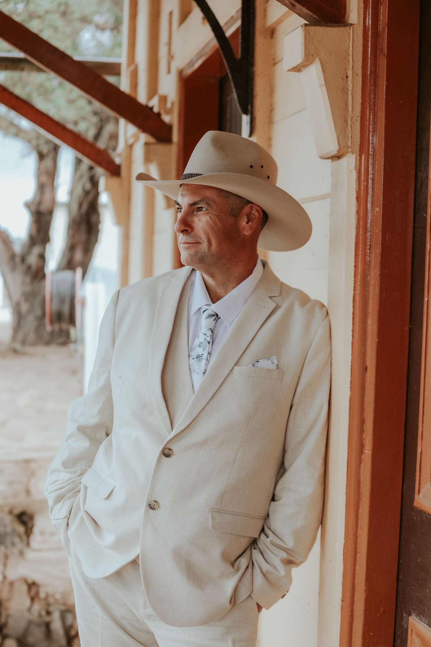 Groom in white suit and cowboy hat standing by wooden doorframe in rustic setting.
