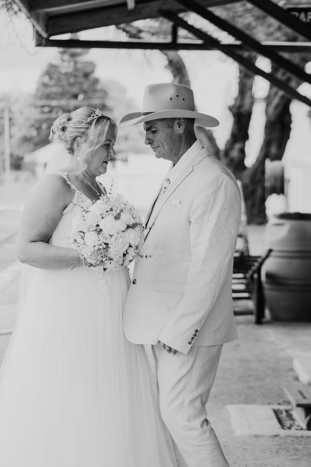 Romantic wedding couple in white attire share tender moments under an outdoor pavilion.