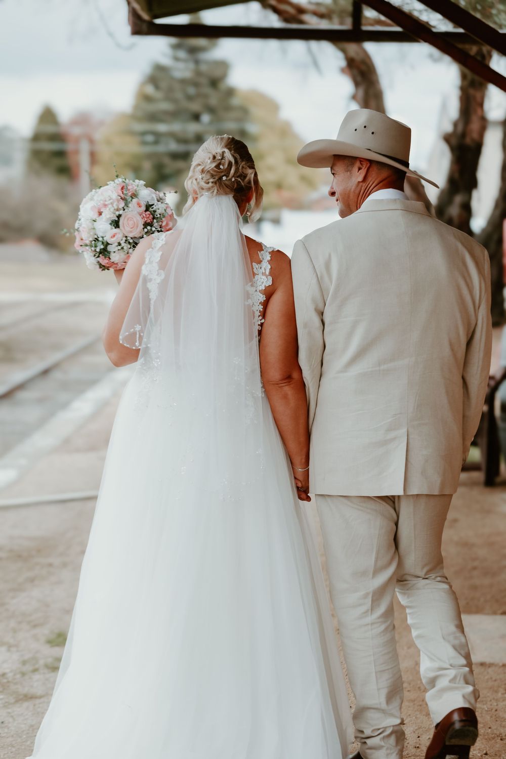 Wedding couple in white and beige attire walk together outdoors under trees.
