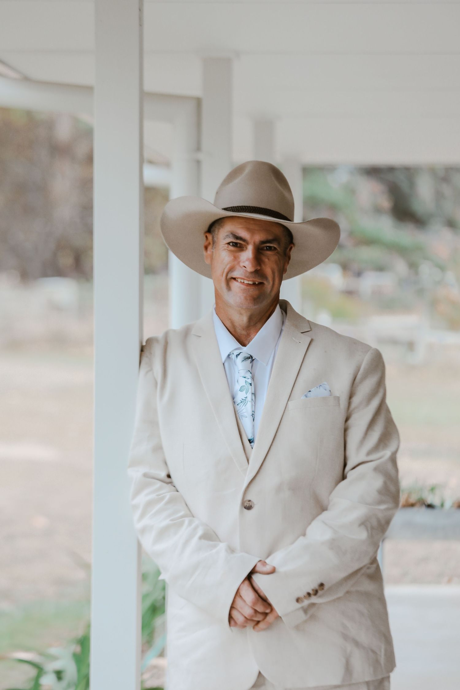 Groom in beige suit and cowboy hat poses under white columns.