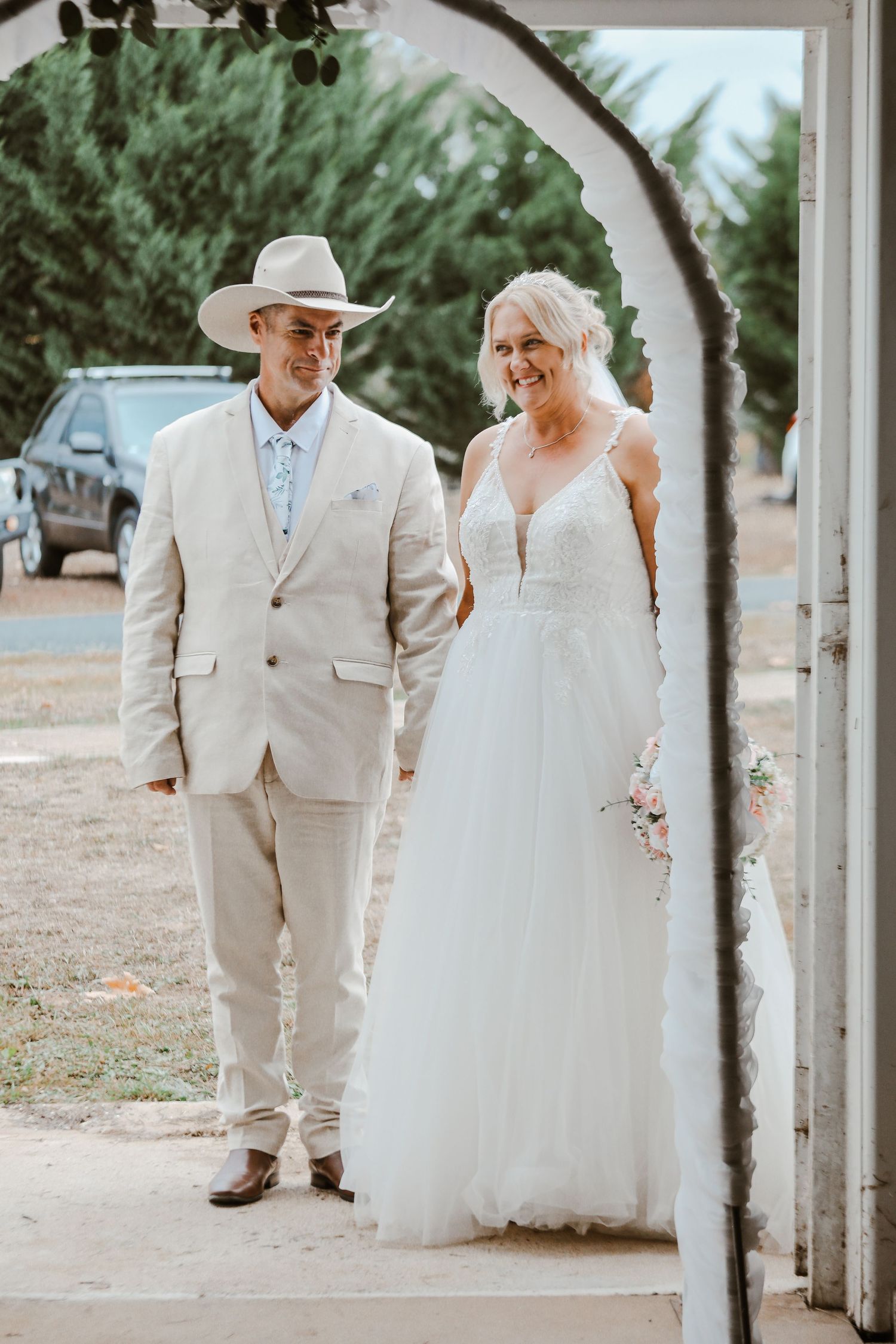 Wedding couple walking under rustic archway in country setting with light colored attire.