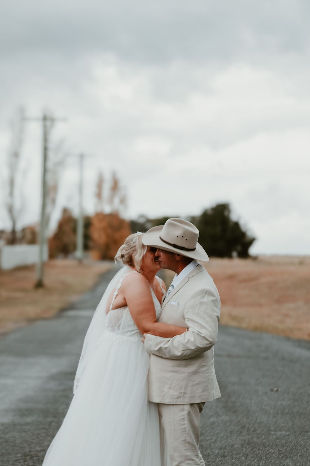 A couple sharing an intimate moment on a cloudy road, the bride wearing a white dress and groom in a light suit and hat.
