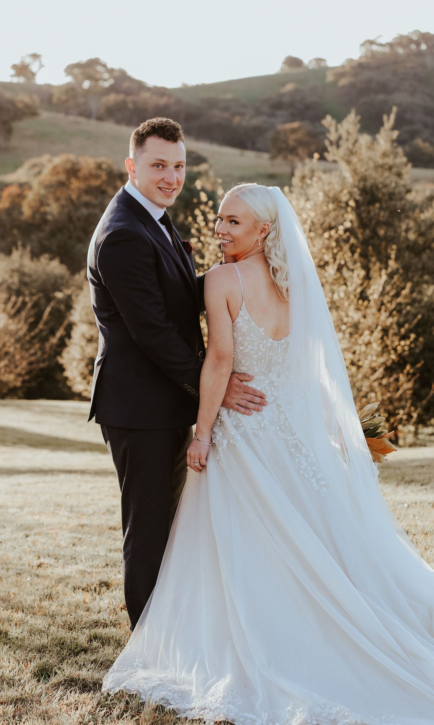 Newlyweds share romantic moment in golden sunlight on grassy hillside.