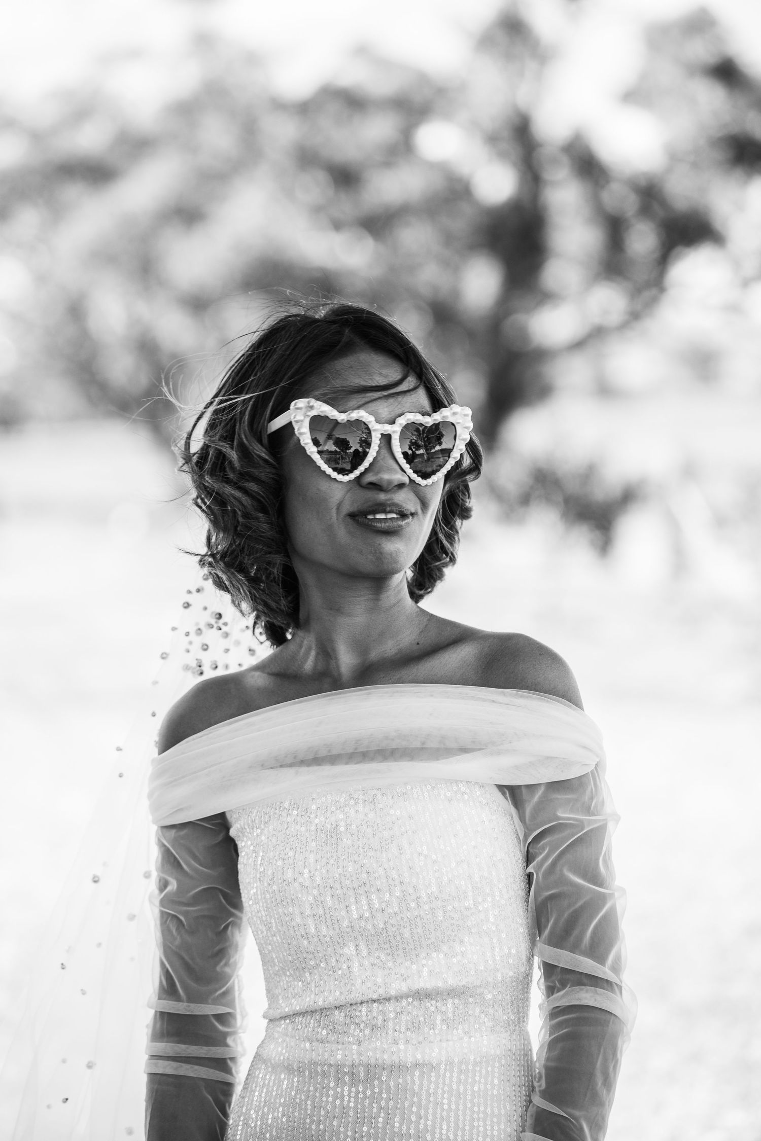 A bride wearing a sparkly off-shoulder dress and heart-shaped sunglasses smiles joyfully.