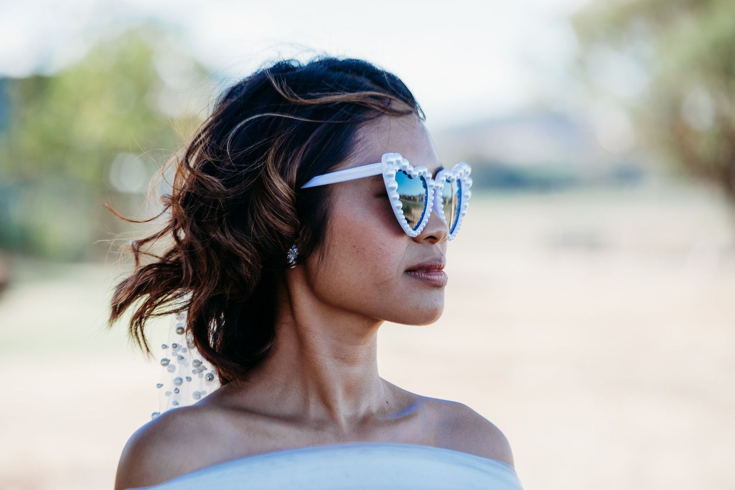 Bride in retro heart sunglasses and off-shoulder dress poses outdoors with a playful expression.