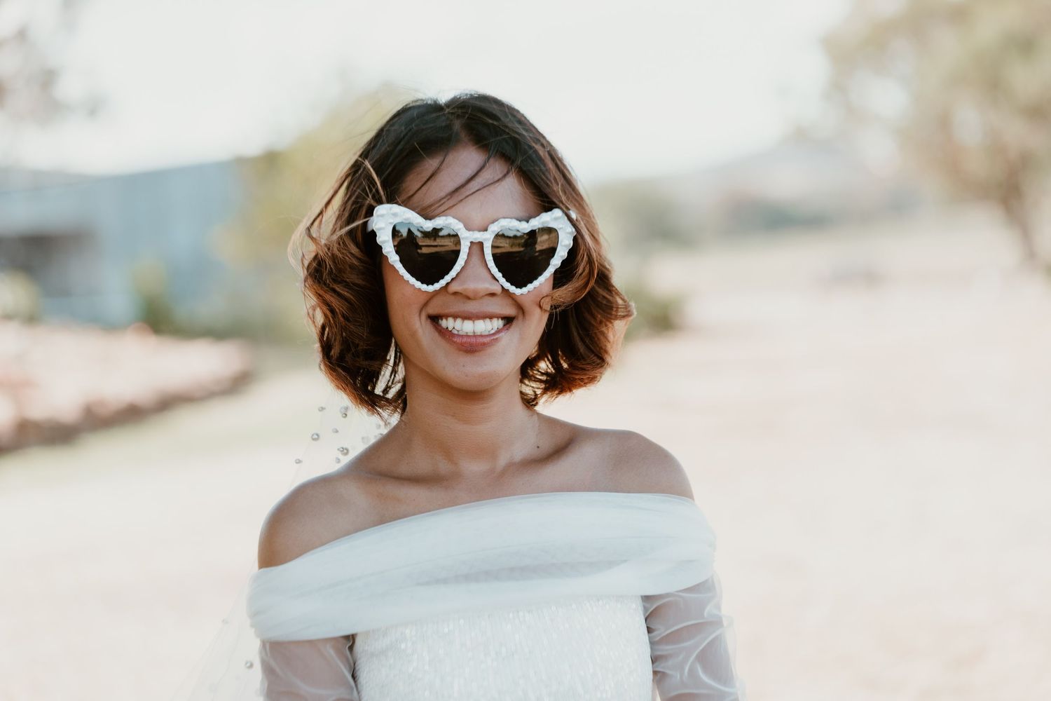 Bride in white dress and heart sunglasses smiles radiantly in outdoor sunlight.