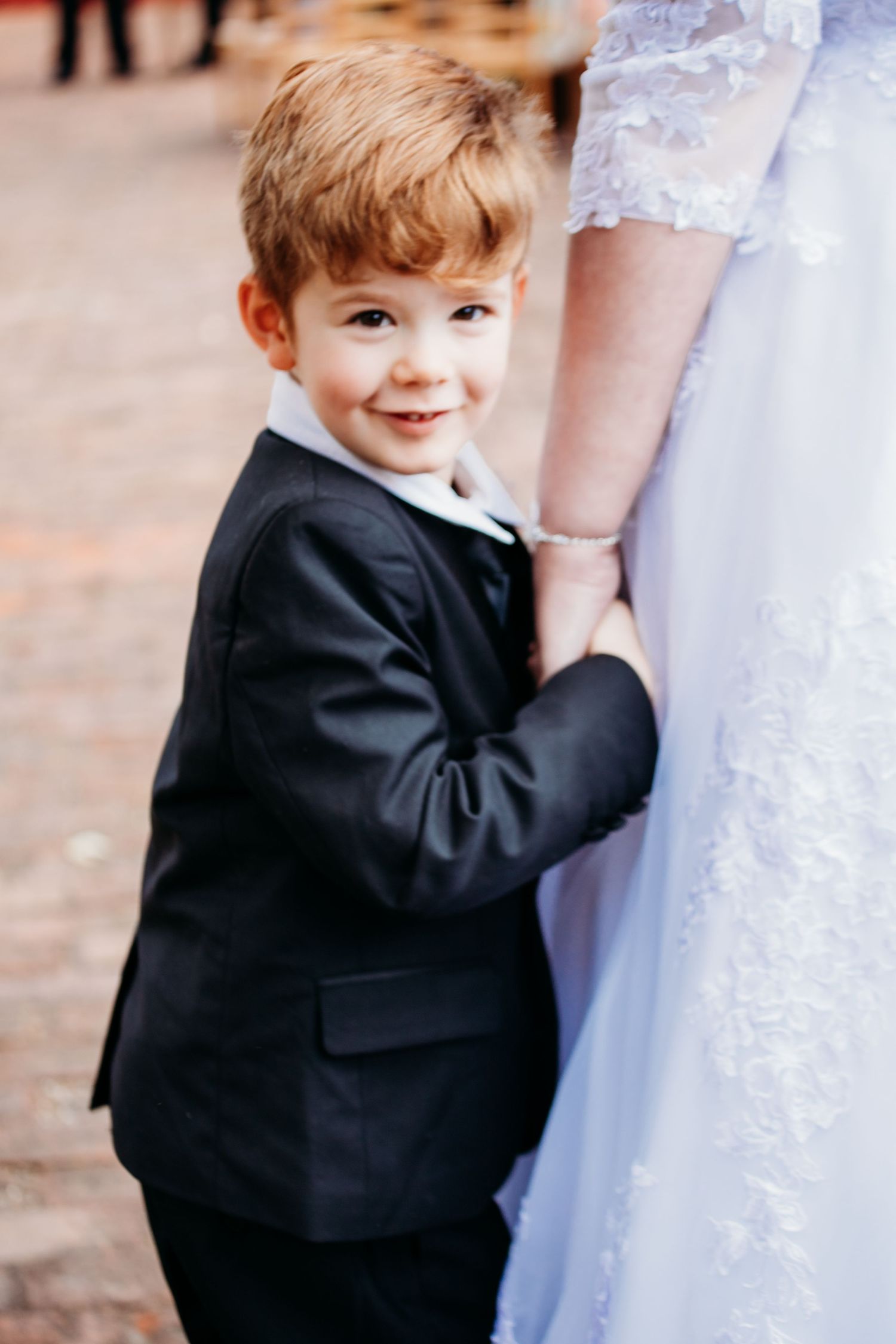 Young ring bearer in black suit plays with the bride's white wedding dress.