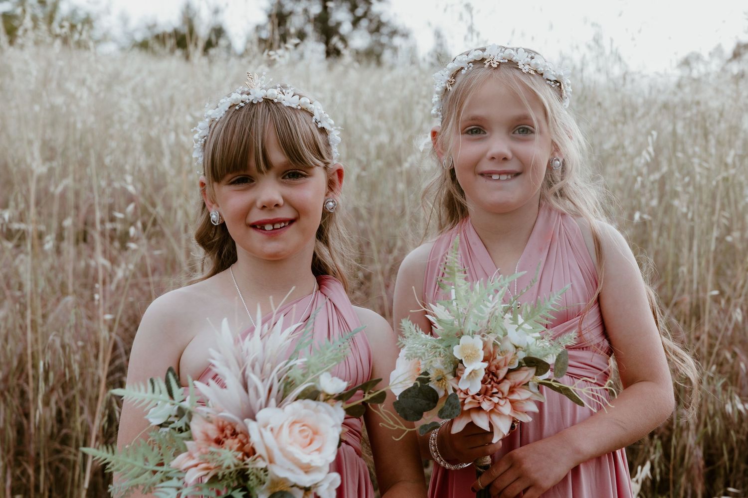 Group wearing pink dresses holds floral bouquets in a rustic outdoor setting.