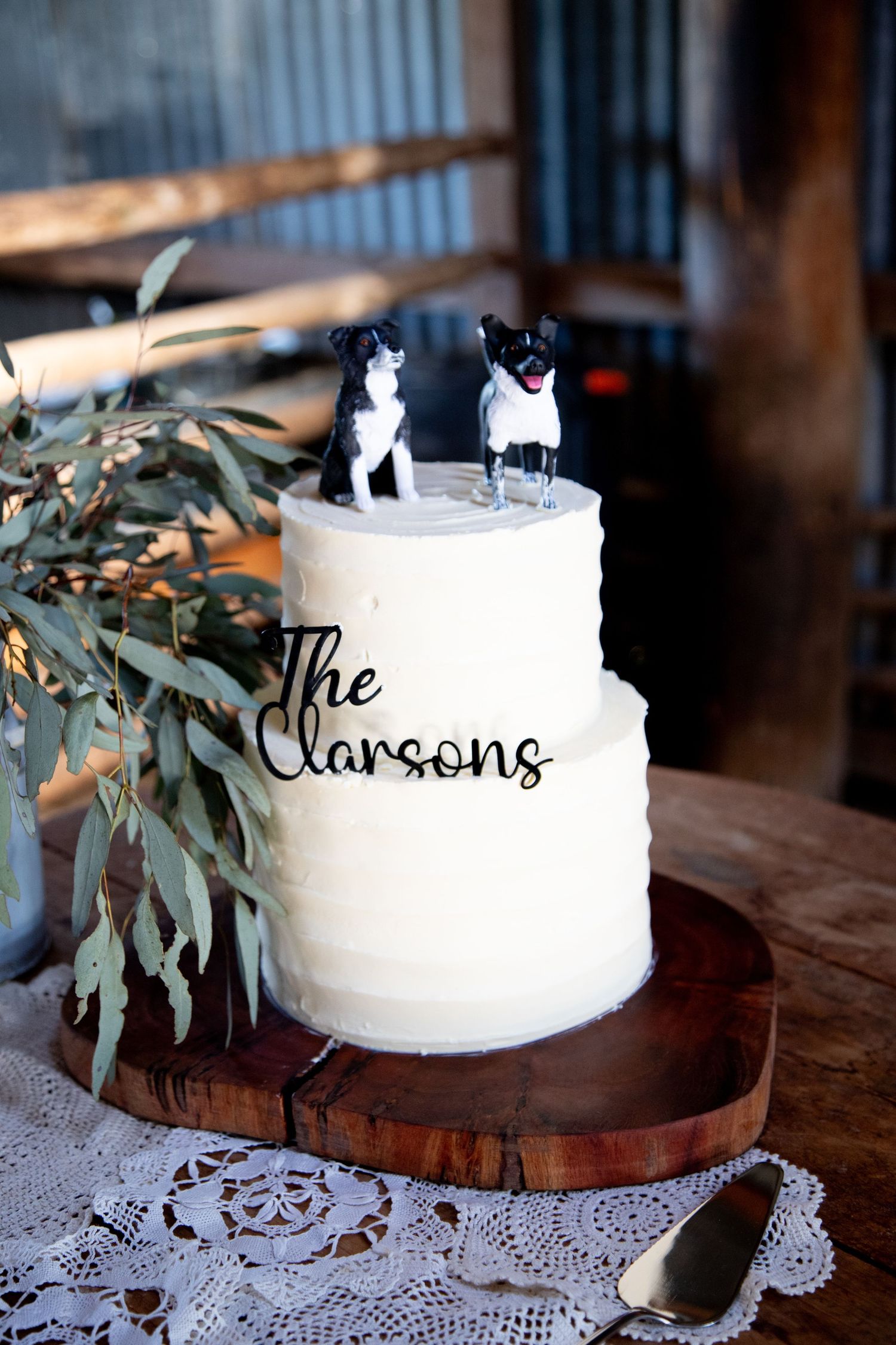 White wedding cake with eucalyptus garnish displayed on wooden stand with lace doily.