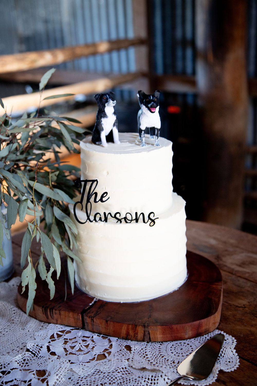 White wedding cake with eucalyptus garnish displayed on wooden stand with lace doily.