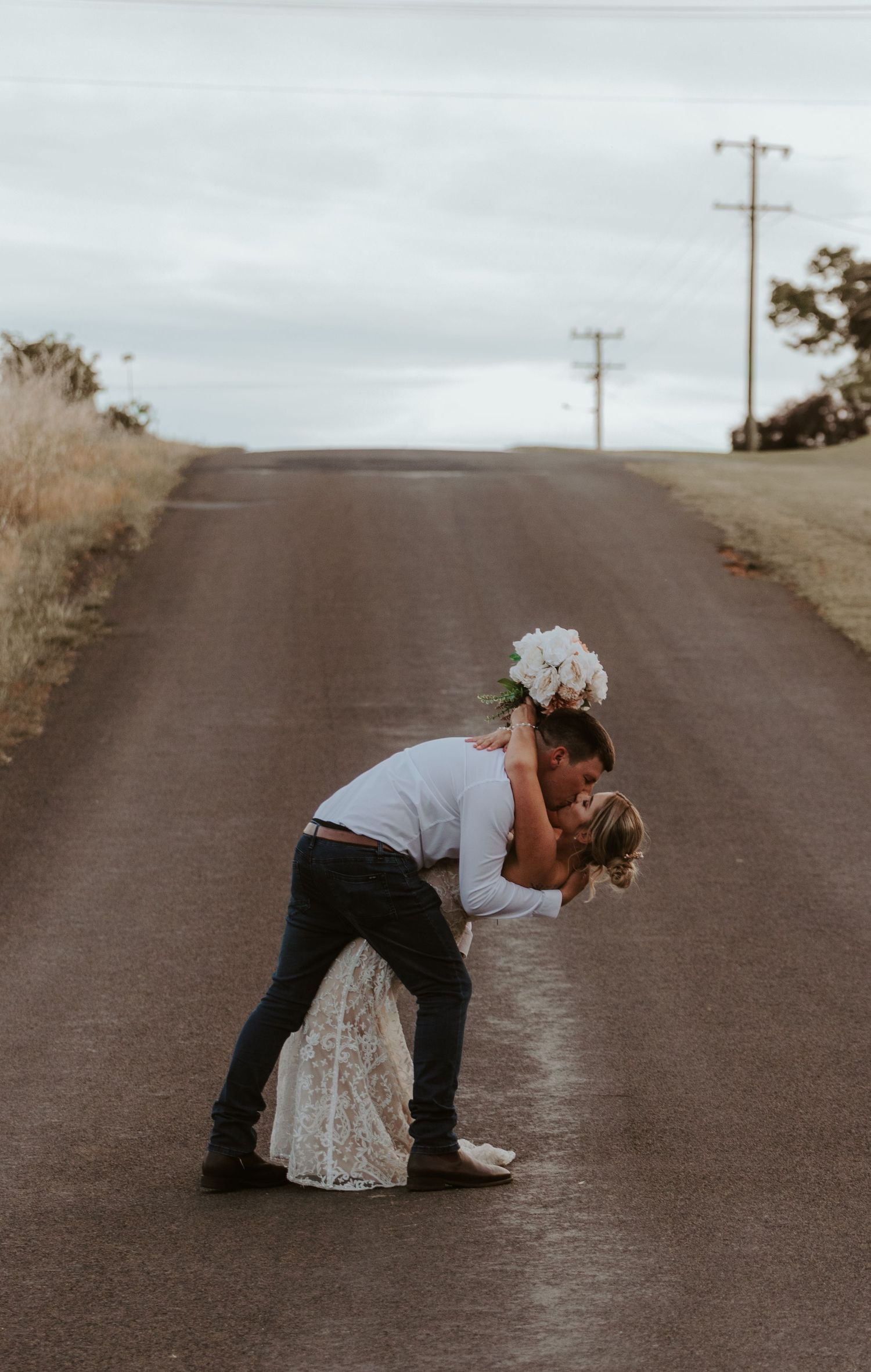 Playful newlyweds pose dramatically on a rural road after their wedding ceremony.