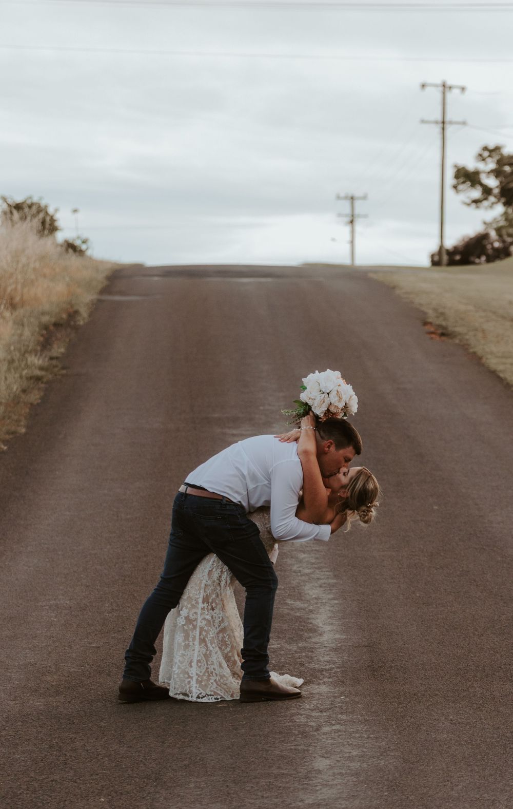 Playful newlyweds pose dramatically on a rural road after their wedding ceremony.
