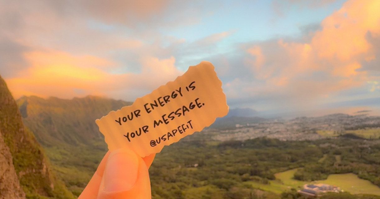 A hand holds up a handwritten note against a scenic mountain landscape at sunset.