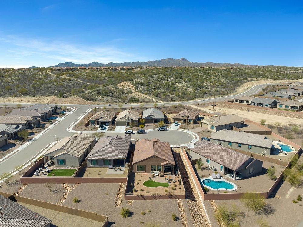 Aerial view of modern desert homes with swimming pools nestled against mountainous terrain under bright blue skies.