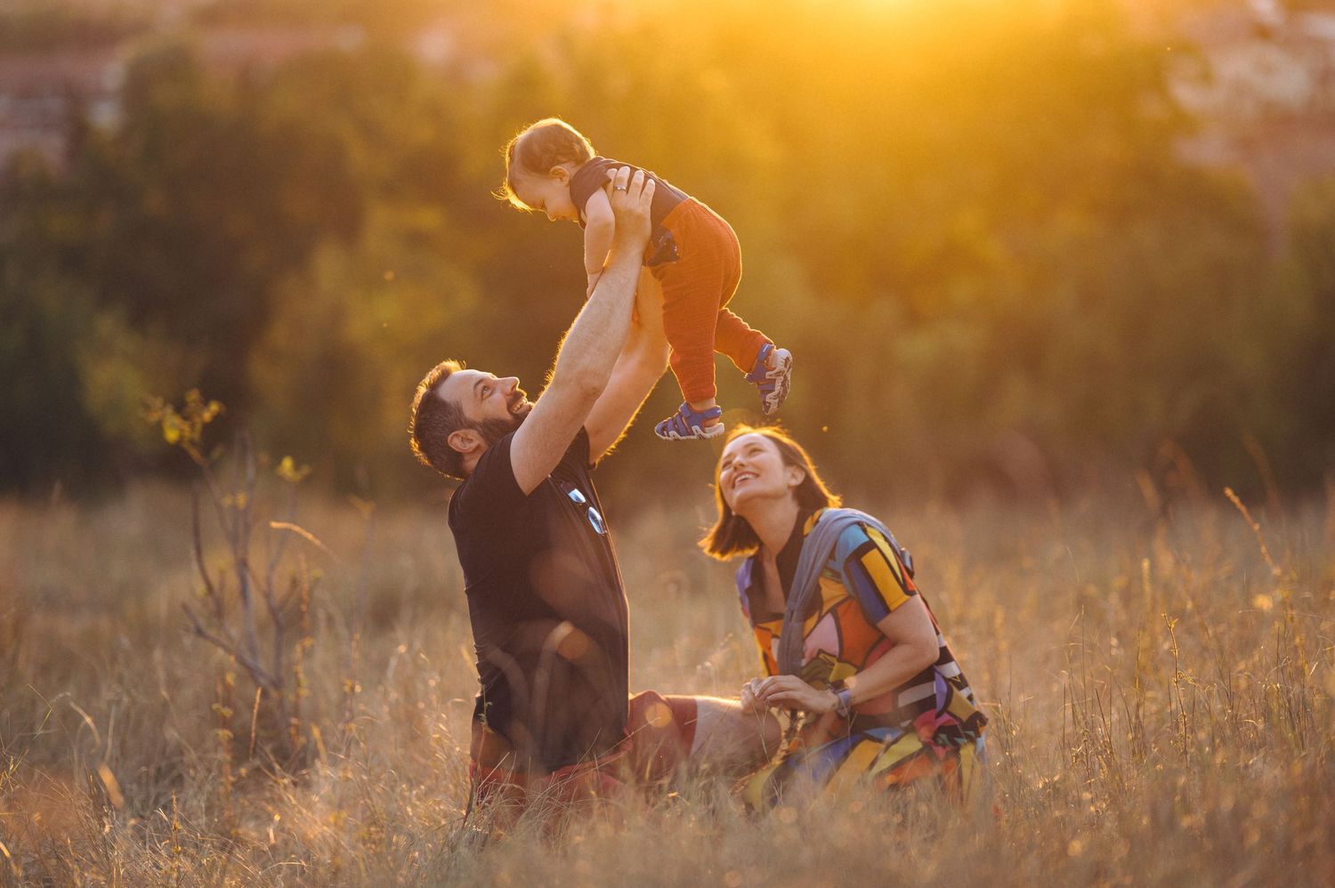 A parent holds a small child up in the golden sunlight while spinning through a grassy meadow at sunset.