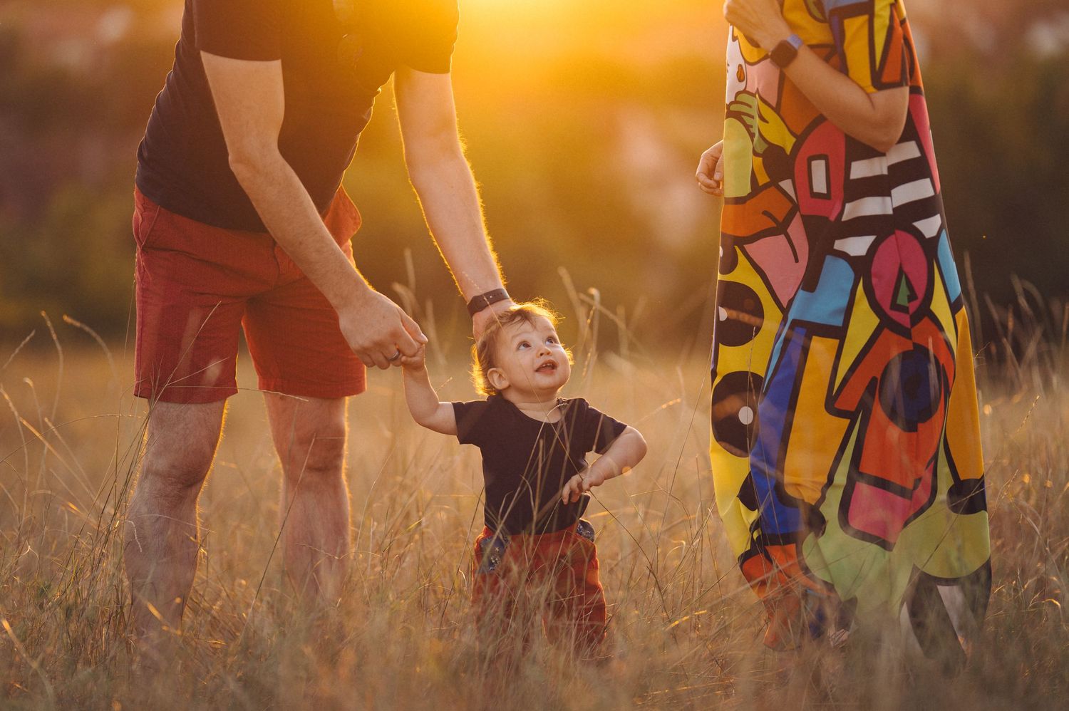A small child stands between two adults in a grassy field during golden hour sunset.