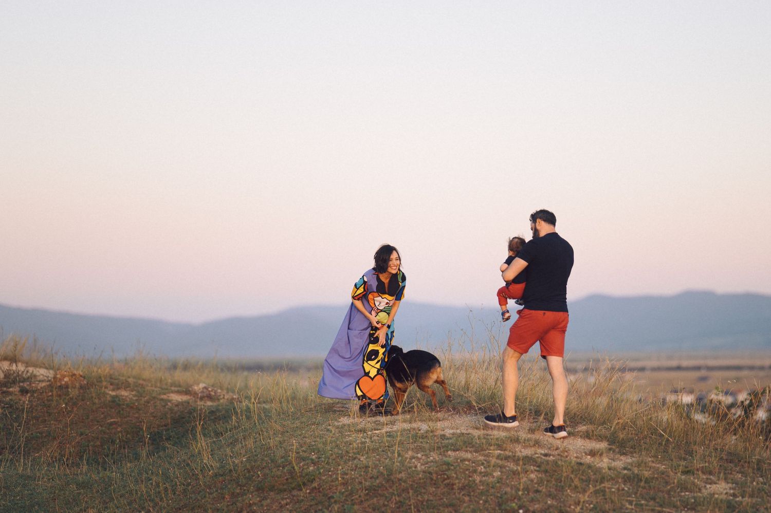Three people sharing a sunset moment on a hilltop with mountains visible in the distant background.