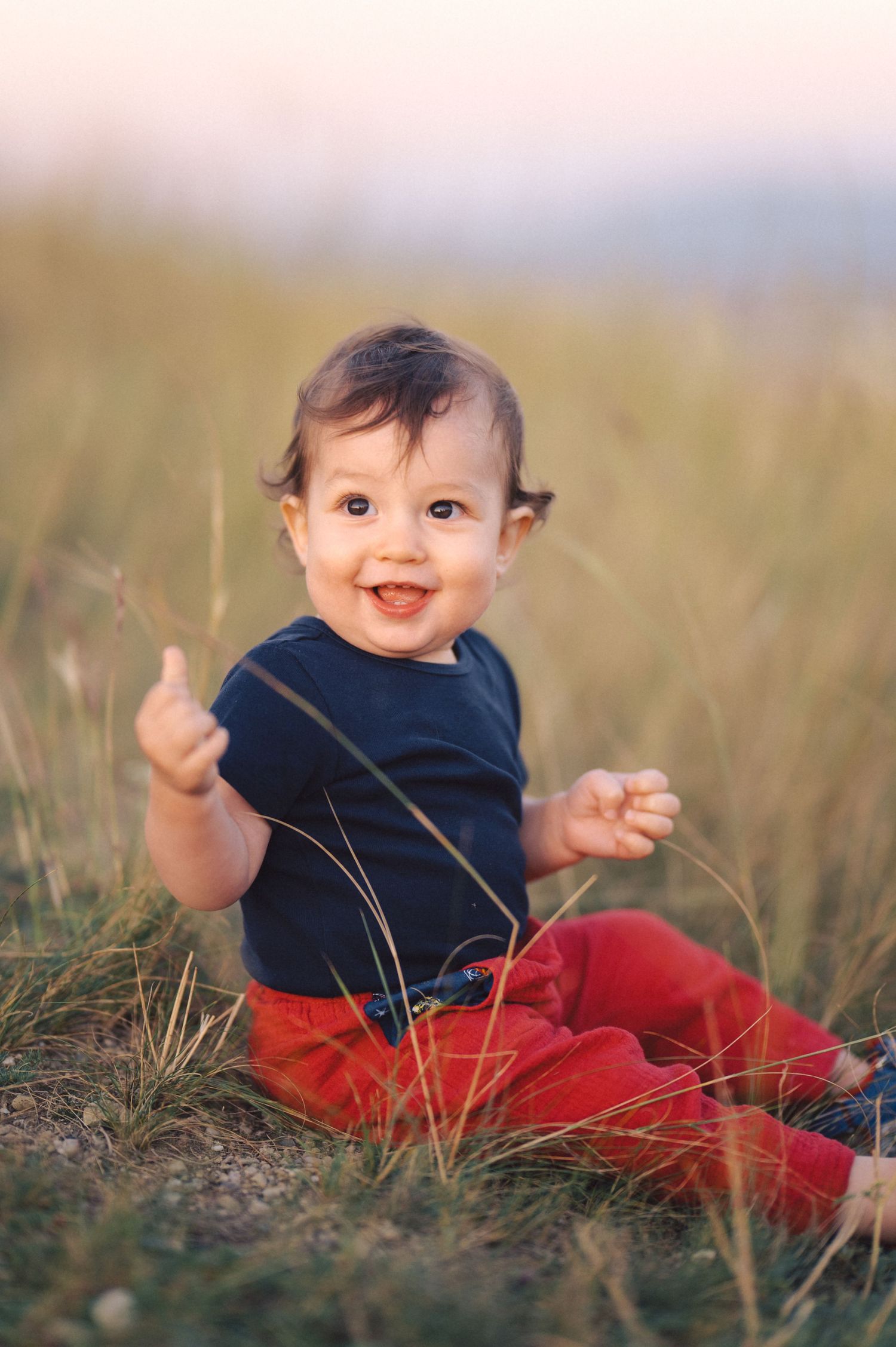 A small child in red pants and navy shirt sits happily in tall grass during a sunny outdoor photo session.