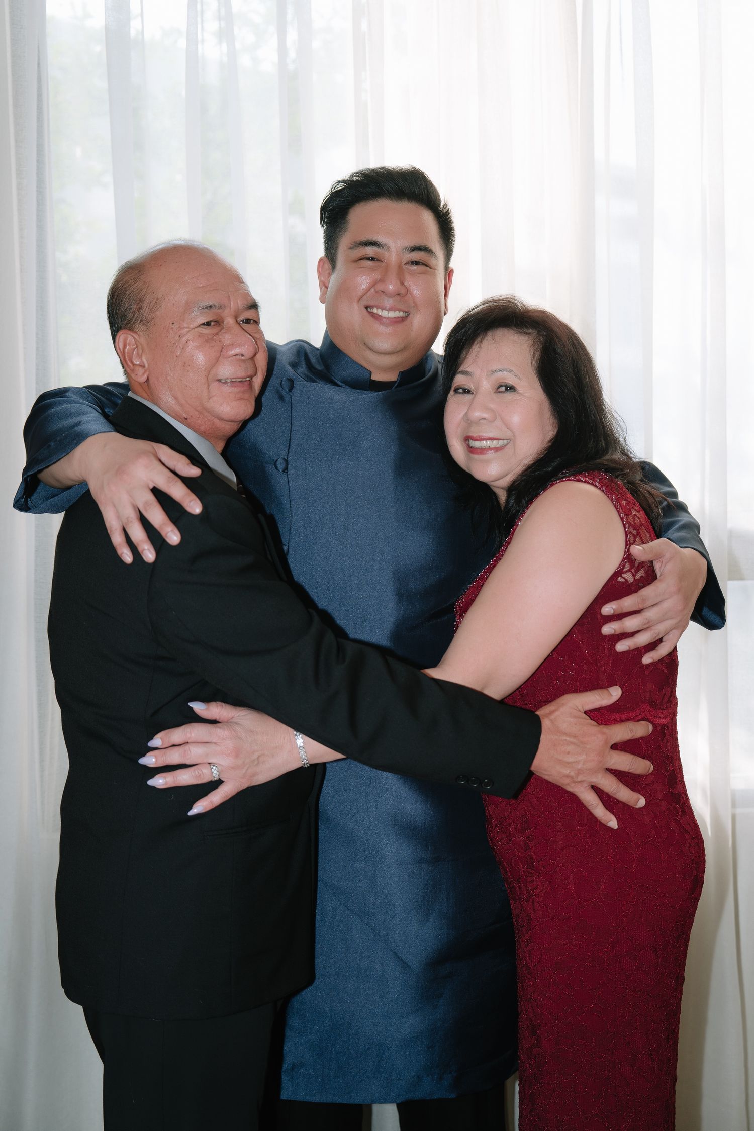 Smiling groom in blue mandarin suit embracing his parents before the wedding ceremony.