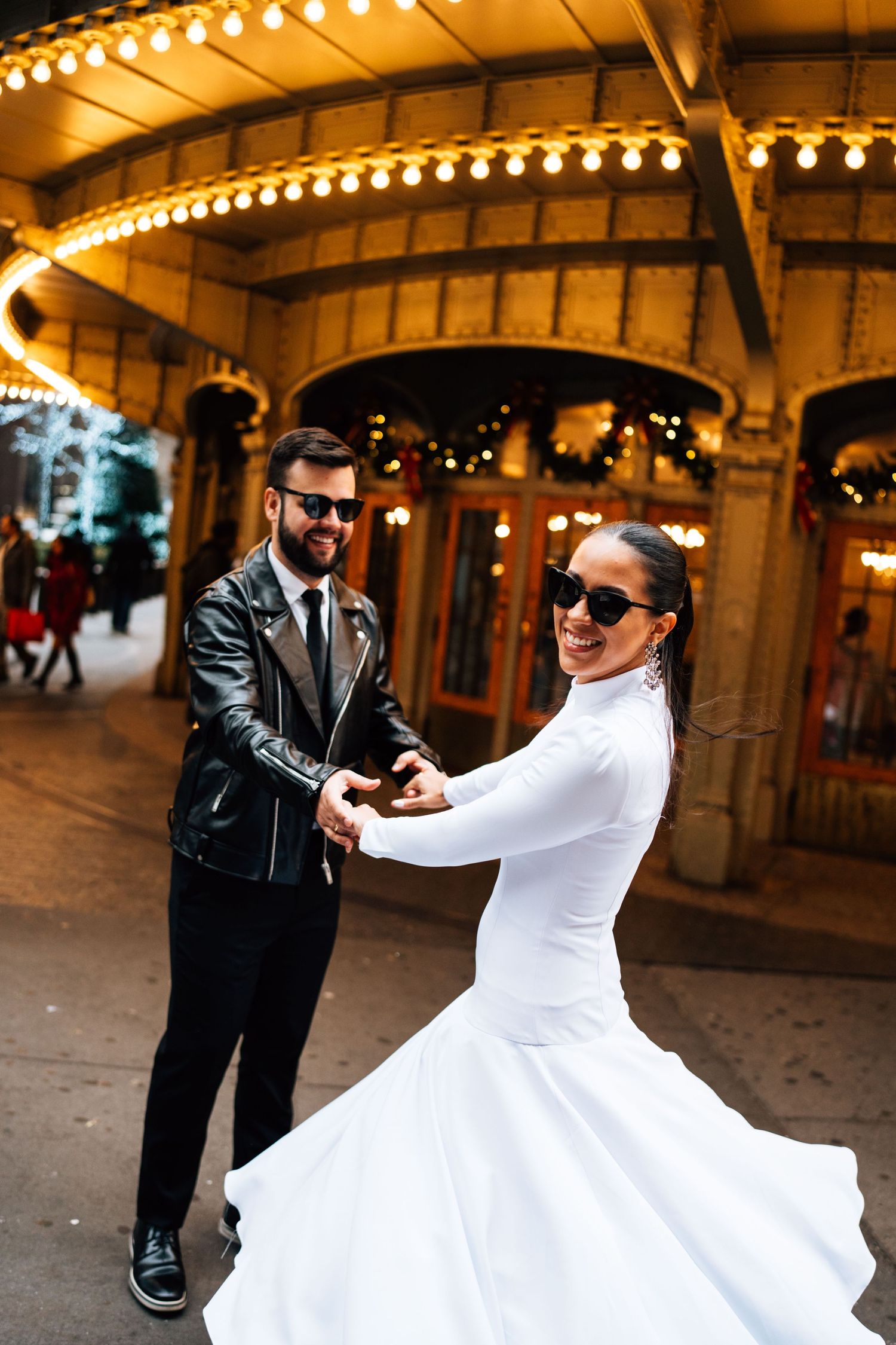A couple in wedding attire spins and dances together beneath warm lights of an urban archway at night.