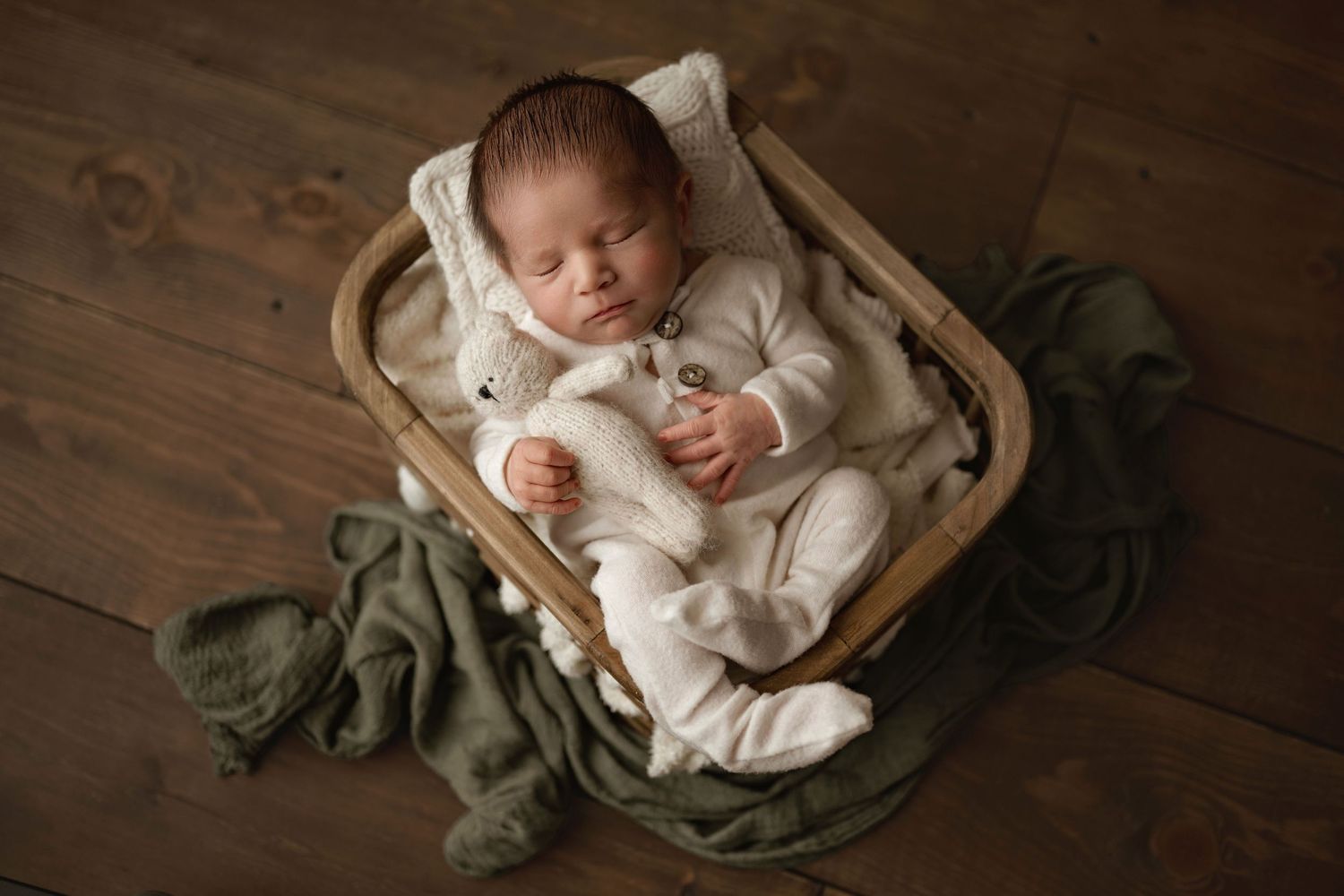 Newborn baby sleeping peacefully in a wooden basket wearing a white romper, surrounded by a green blanket on hardwood floors.