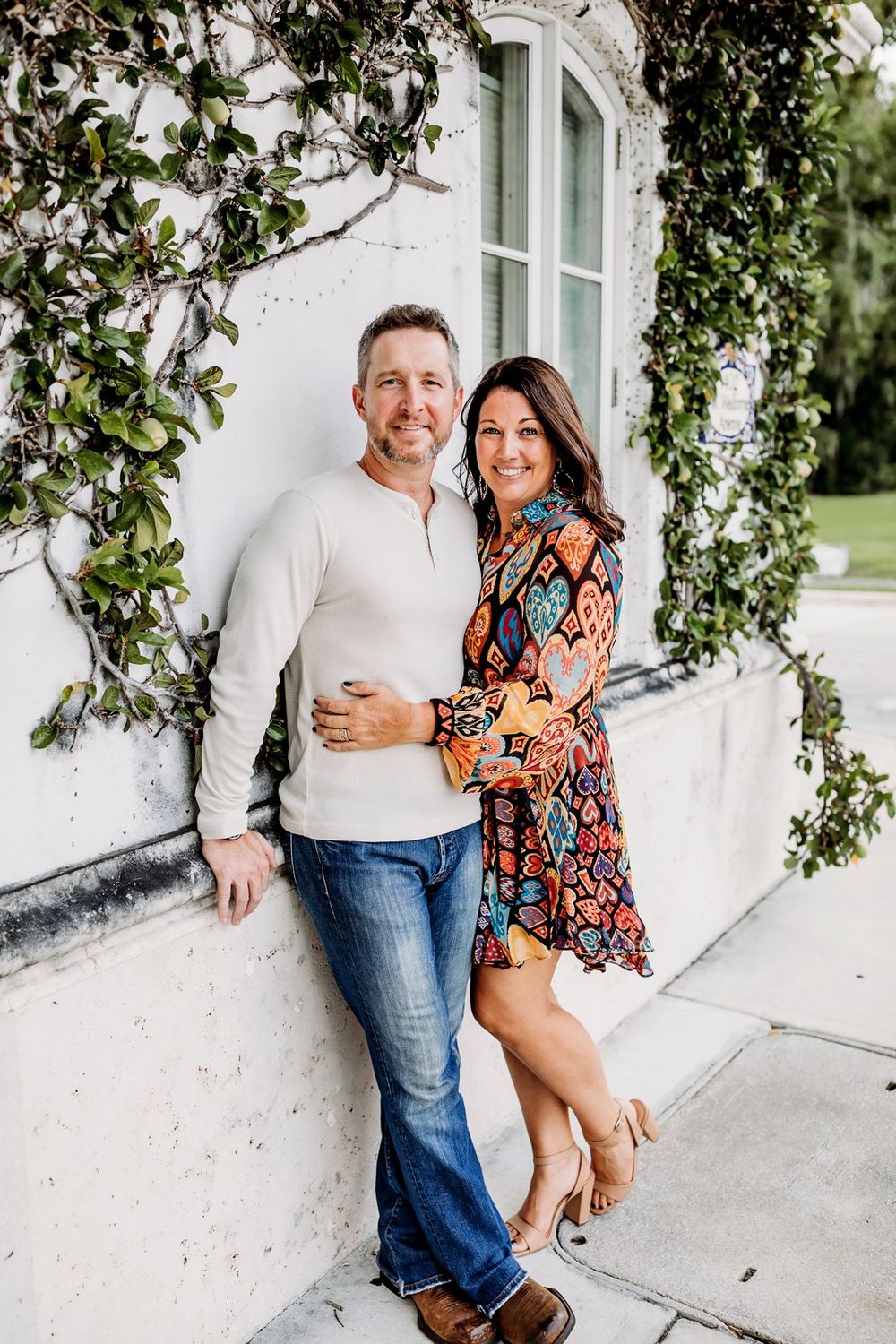 Couple poses lovingly together on white covered porch with lush green foliage in background.