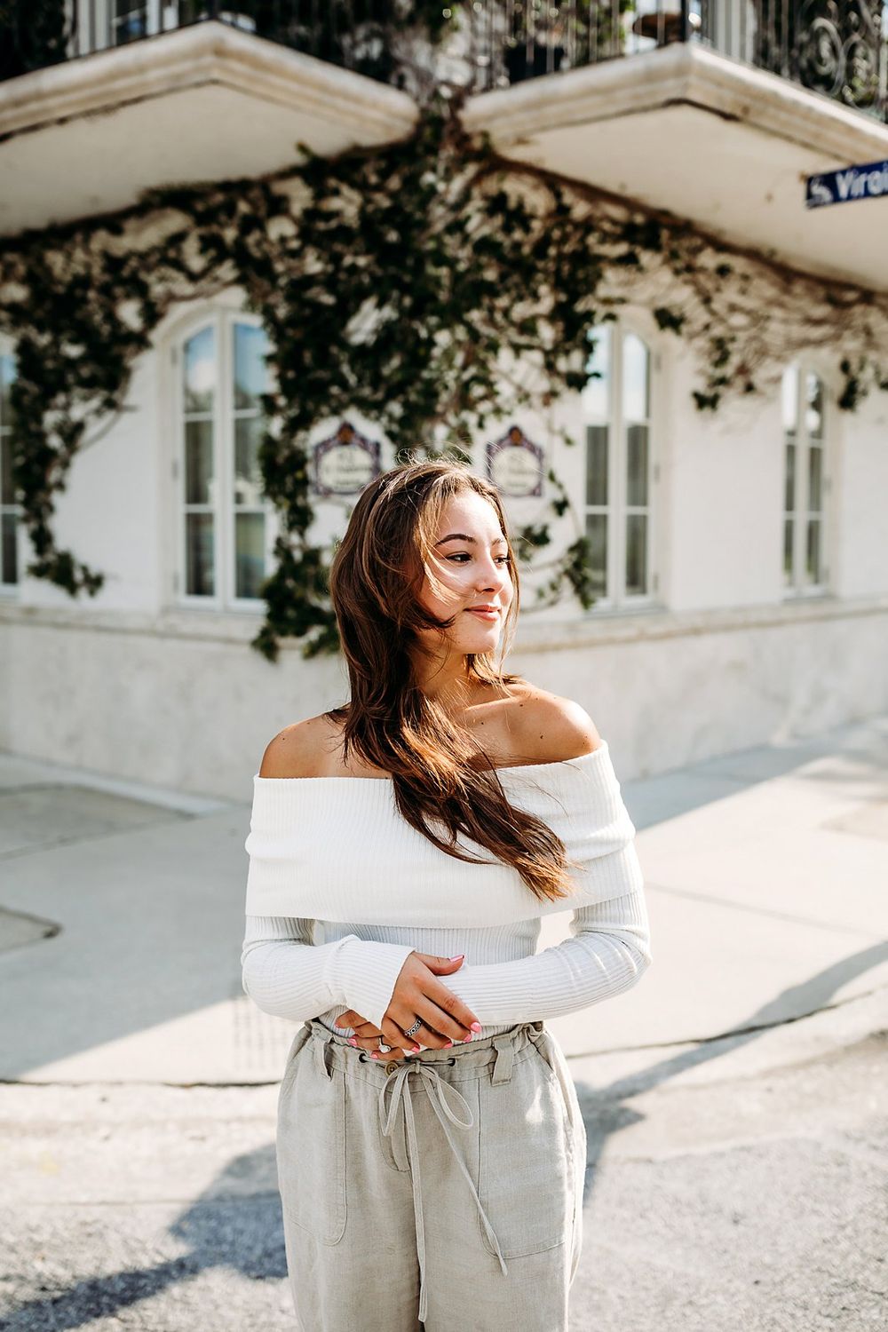 Woman in off-shoulder white top and light pants enjoying sunny day by elegant building with white facade.