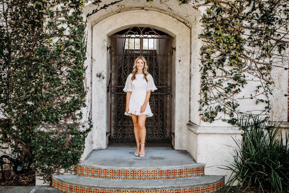 A person in a white dress stands in front of an elegant stone archway entrance surrounded by lush greenery.