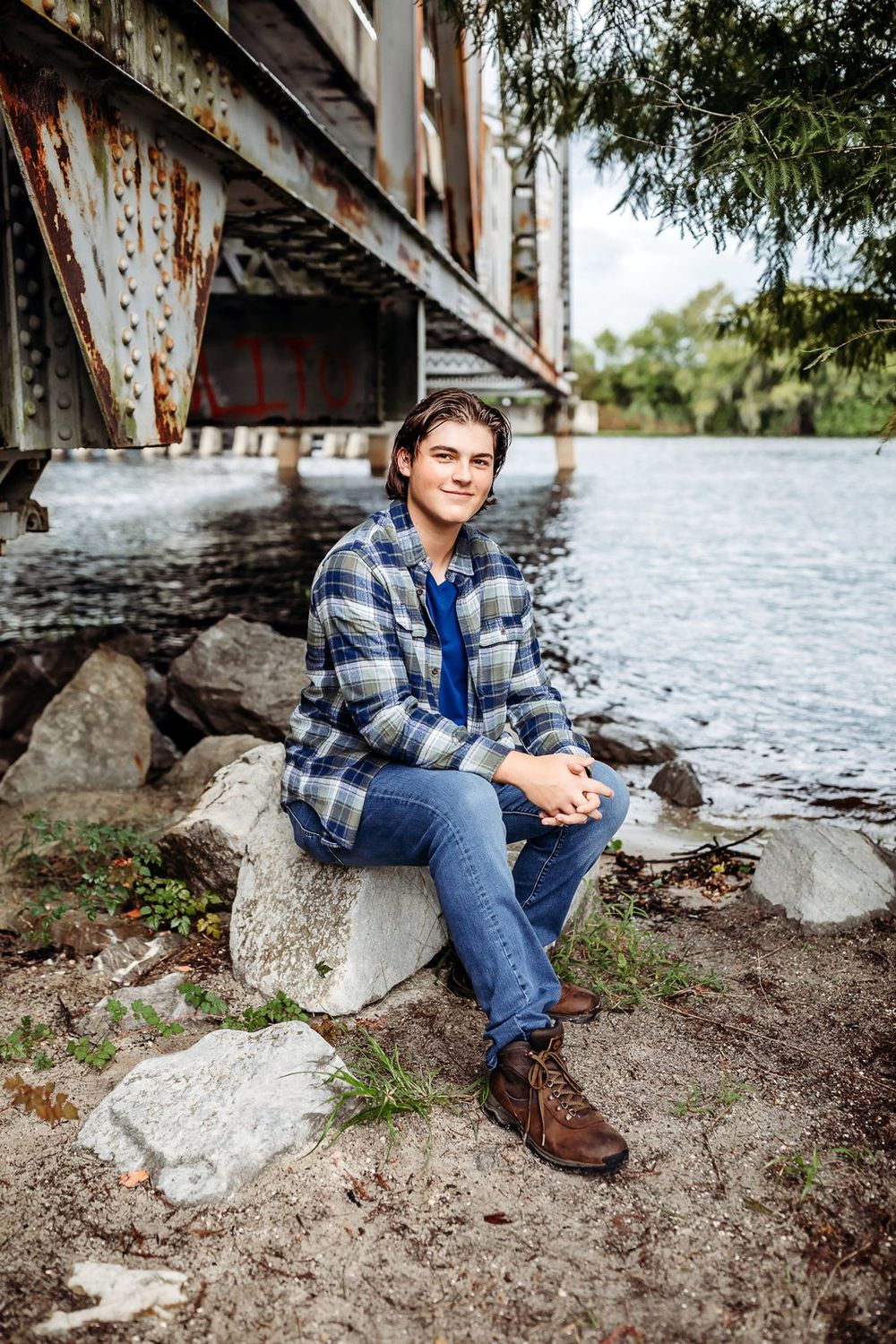 Subject sits casually on rocky riverbank wearing plaid flannel shirt and jeans in outdoor portrait series.