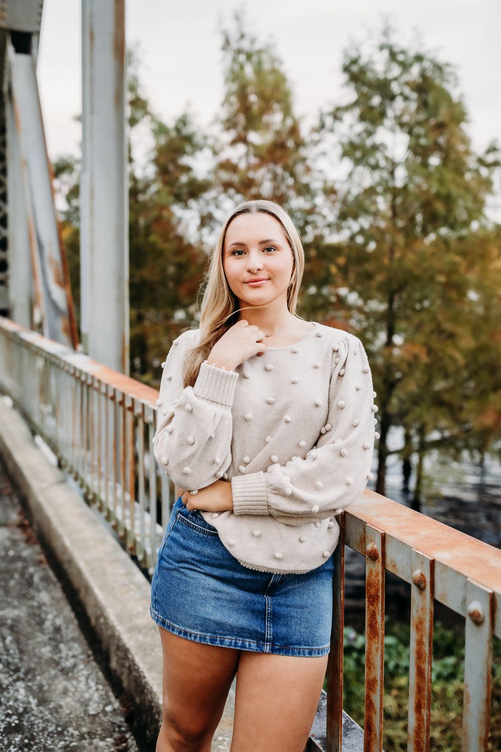 Woman in cozy beige sweater and denim shorts poses against outdoor railing in autumn setting.