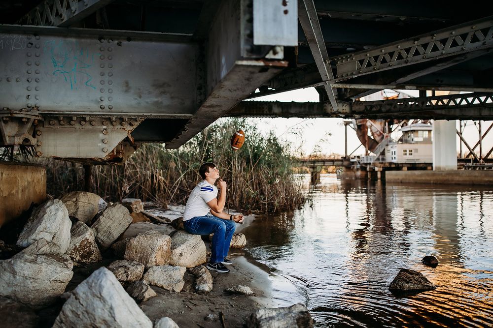 Person sitting on rocks under a rustic bridge beside calm water at sunset.