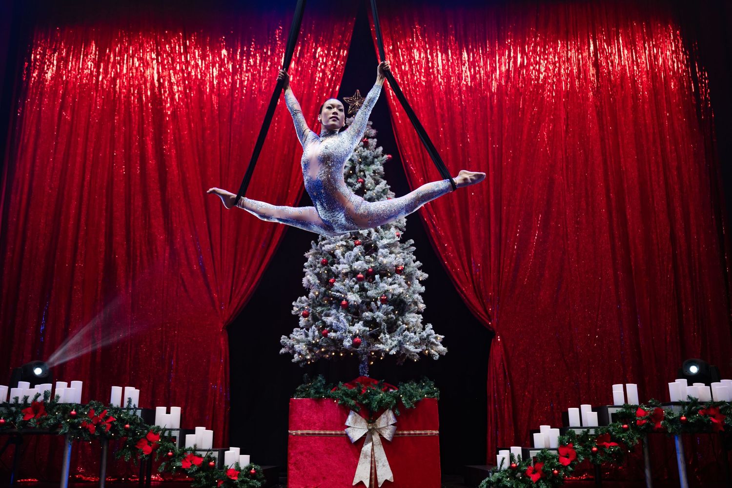 Performer in sparkling silver costume strikes dramatic pose against red velvet curtain backdrop during burlesque show.