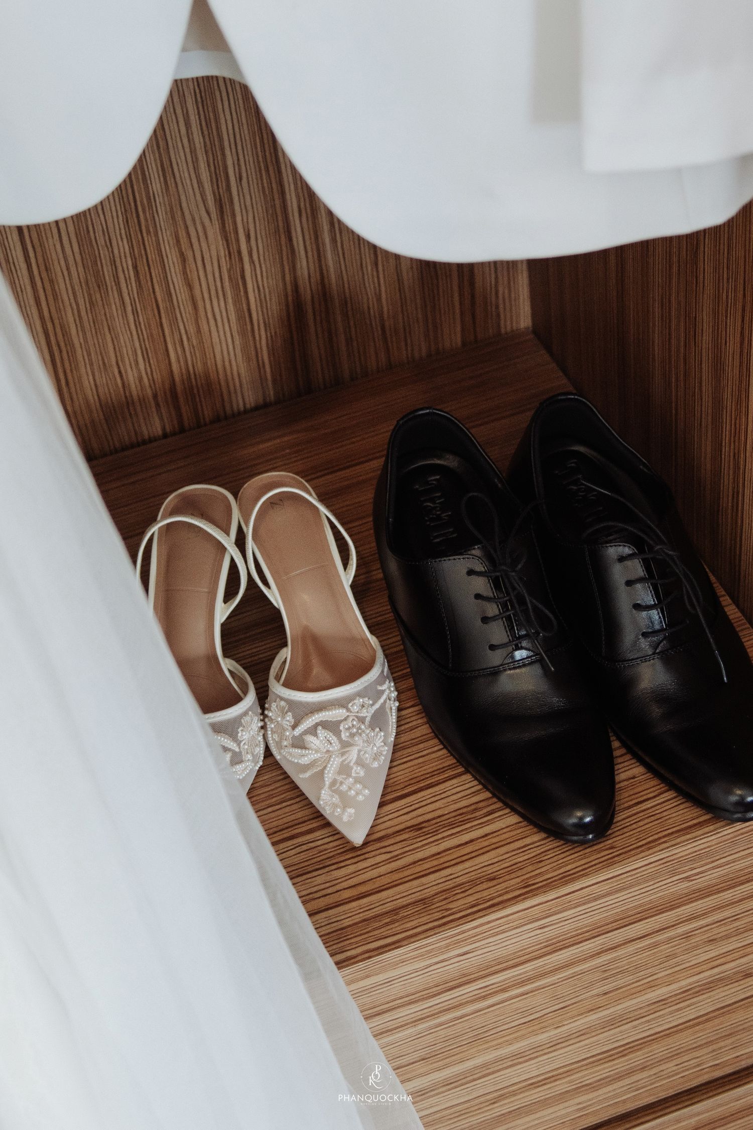 Bride white embellished slingback heels and groom black Oxford dress shoes displayed on wooden shelf together.