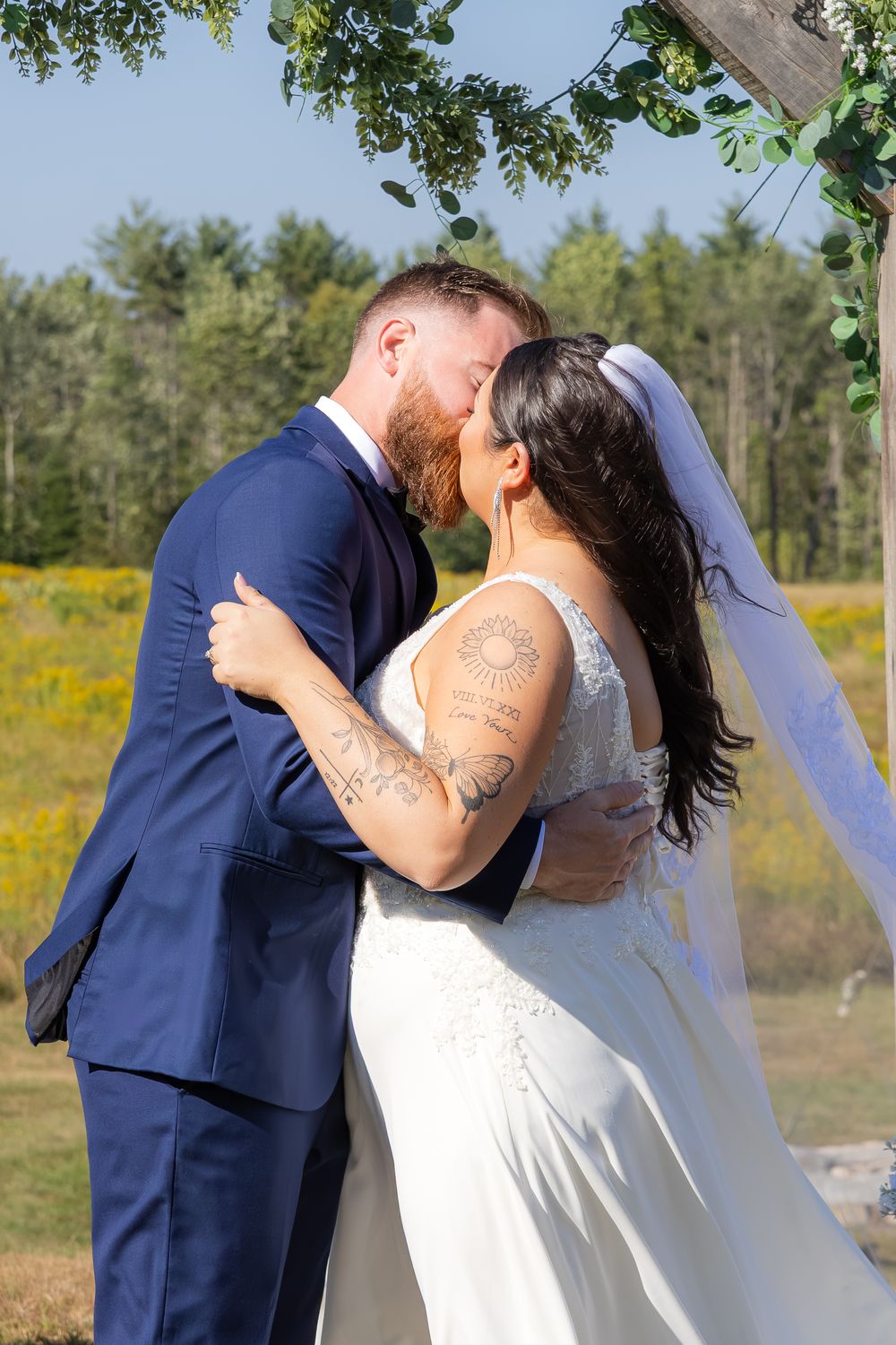 A newly married couple embraces and kisses in a sunlit outdoor setting with trees and yellow flowers in background.