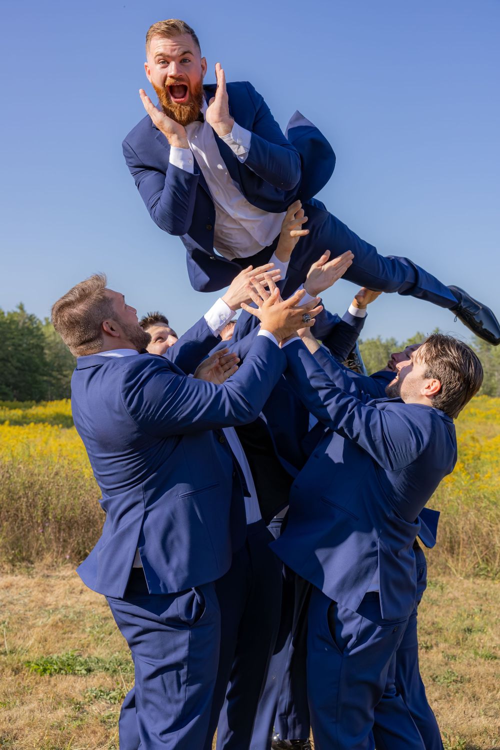 Two people in navy blue suits playfully interact during a photoshoot in a sunny field with yellow flowers.