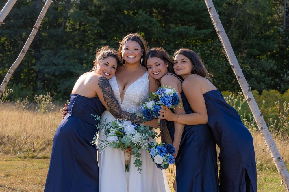 A bride and bridesmaids in navy blue dresses pose together outdoors on a sunny day.