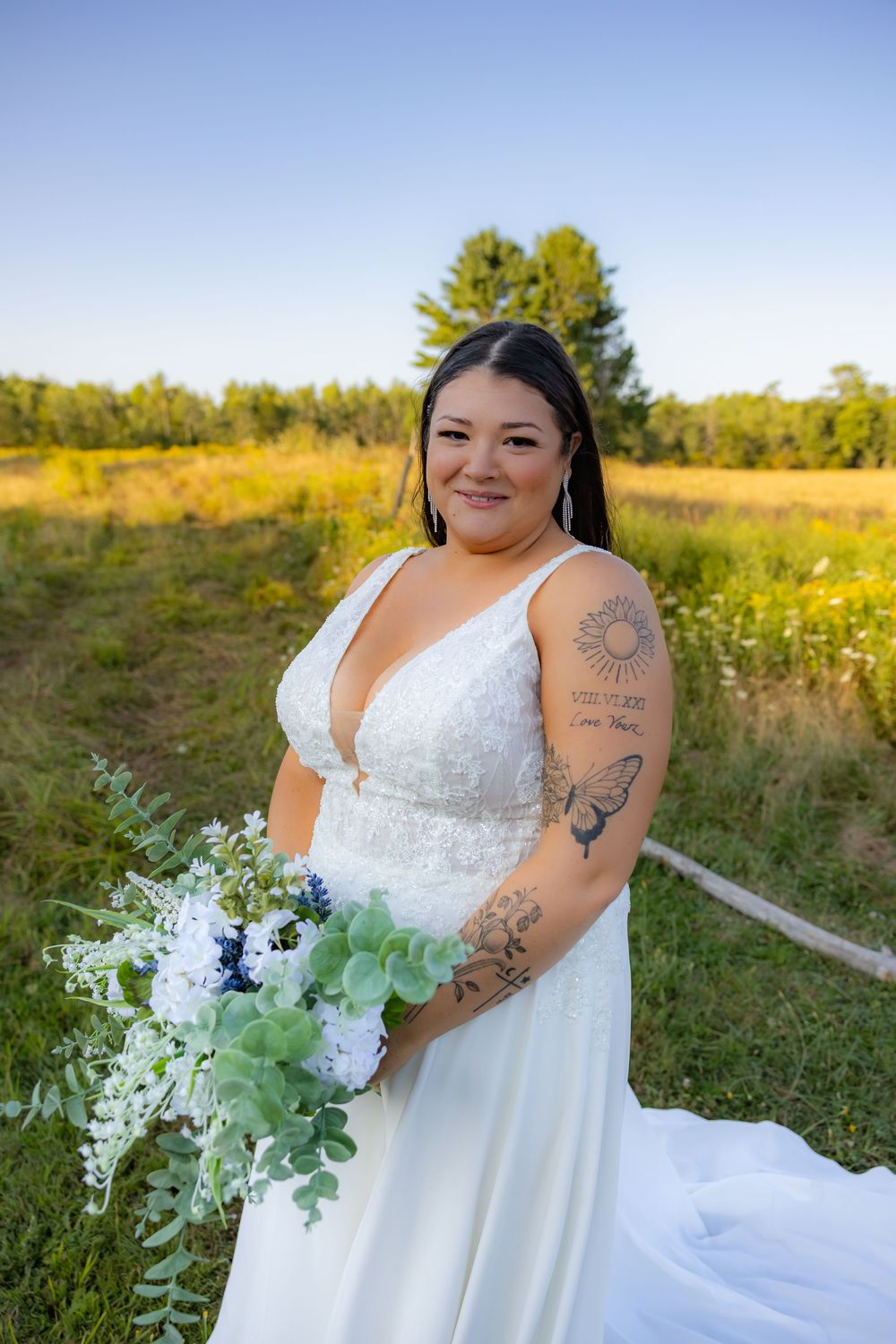 A bride in a flowing white wedding dress poses in a golden field during sunset holding a bouquet with greenery and flowers.